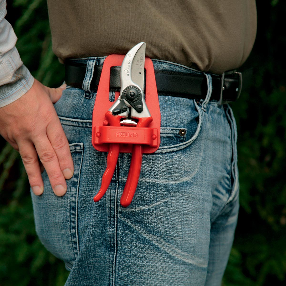 Freund Beskæresaks 2002 i rødt bælteetui på jeans; smedet aluminiumskrop, udskiftelig kulstofstål klinge, trådskærer, riflet bolt og støddæmper – professionel og holdbar beskæresaks fra Freund Victoria