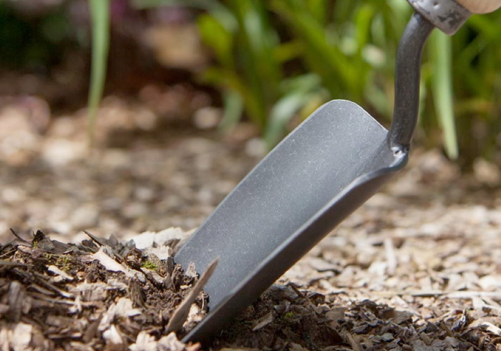 Nærbillede af en metal håndspade, som graver i barkflis i en have