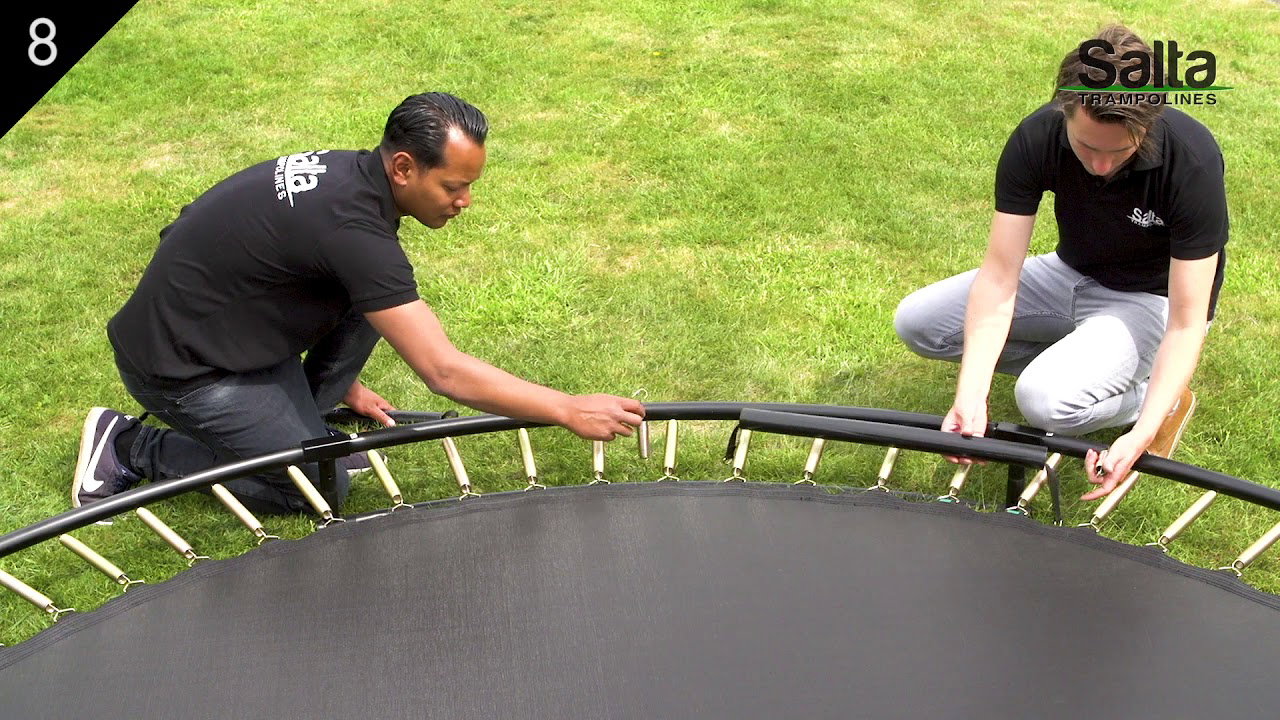 To personer monterer Salta Trampolin Royal Baseground Sport Ø305 cm (sort) på græs; synlige fjedre, sort kantpolstring og sorte rammedele set tæt på hoppefeltet