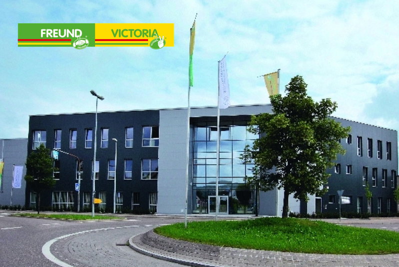 Moderne erhvervsbygning med glasfacade og flag ved rundkørsel Fasade af en moderne treetagers kontorbygning med glasindgang, flere vinduer, flagstænger og et træ ved en rundkørsel; 'FREUND VICTORIA' logo i øverste venstre hjørne.