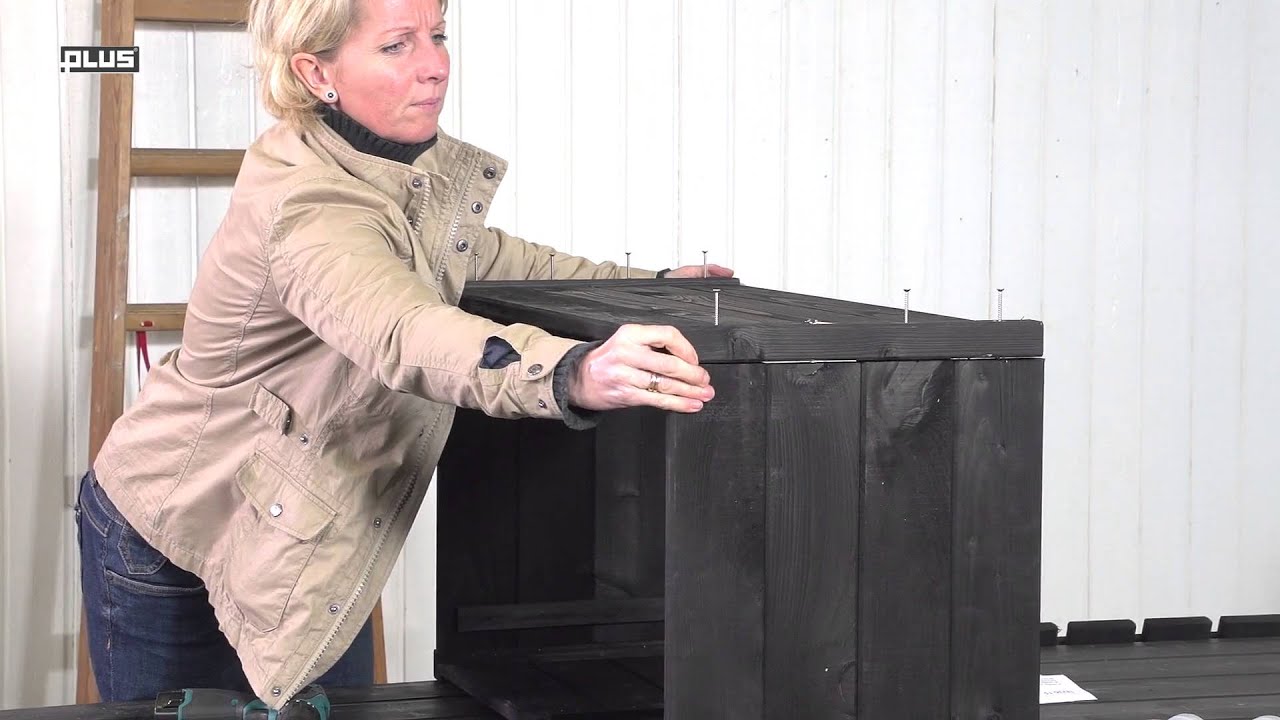Person monterer grundmalet sort træblomsterkasse 48x88x47 cm fra PLUS, vist under samling på arbejdsbord. Robust kapilærkasse til terrasse, altan eller drivhus, perfekt til krydderurter og jordbær, inkluderer hjul.