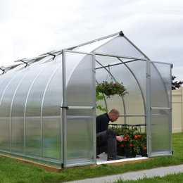 Drivhus Bella 12 m² sølv Palram - Canopia | 2,4 m bredt klokkeformet drivhus med alu-ramme og dobbeltvæggede polycarbonatpaneler, galvaniseret stålbase Drivhus Bella 12 m² i sølv fra Palram - Canopia med åbne dobbeltdøre, klokkeformet design, stærk aluminiumramme og dobbeltvæggede polycarbonatpaneler. Mand plejer tomatplanter indvendig, synlige ventilationsvinduer på taget.