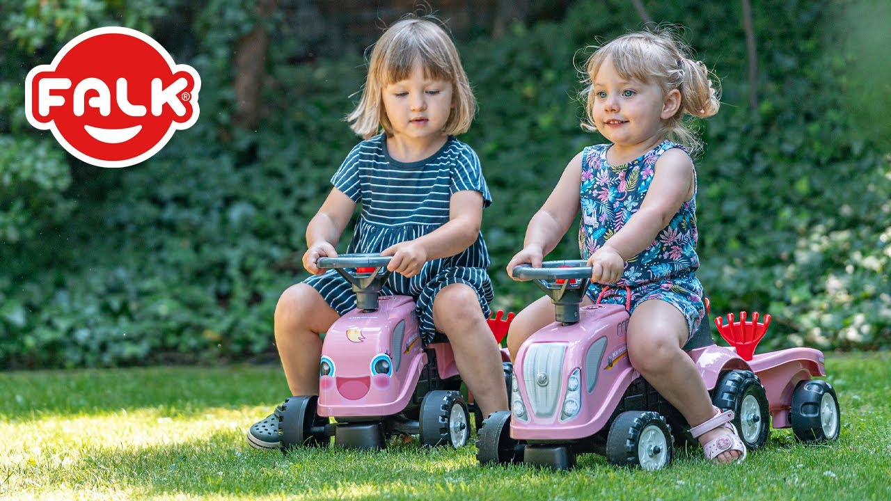 To små piger kører på lyserøde New Holland pedaltraktorer med trailer, rive og skovl i haven. FALK børnetraktor i pink, ideel til børn fra 1 år, drejbart rat og manuelt horn - perfekt til leg og transport af legetøj eller jord.