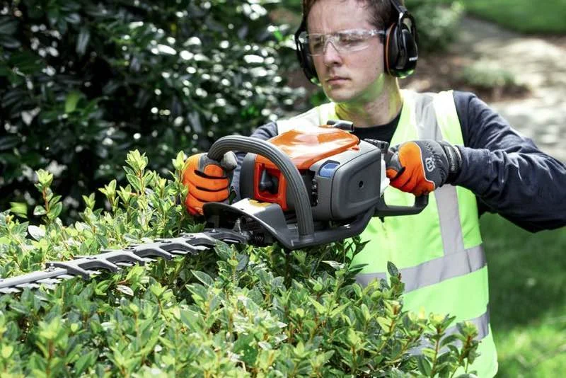 Person iført sikkerhedsbriller, høreværn og refleksvest klipper en hæk med en batteridrevet hækkeklipper