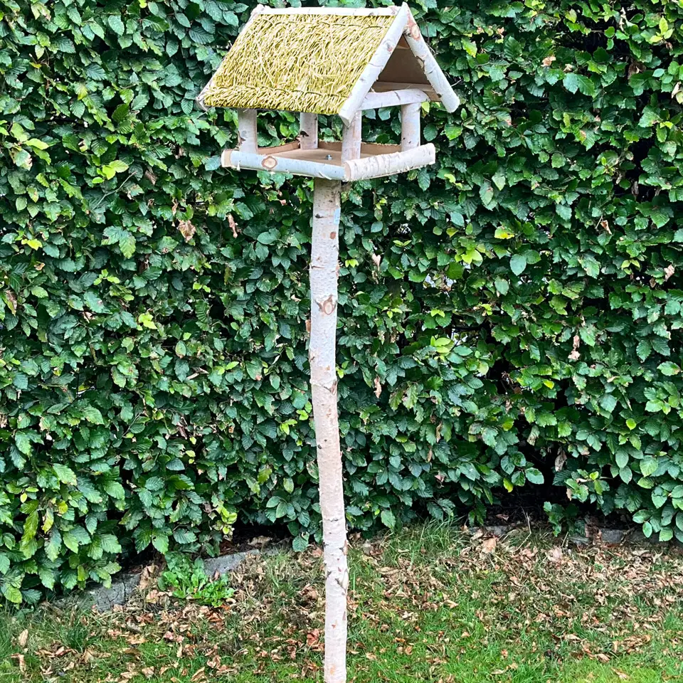 Fuglefoderhus i birketræ på stander med stråtag – HORTUS, 146 cm Fuglefoderhus i ægte birketræ med stråtag på stander (146 cm) fra HORTUS — rummeligt fuglehus hævet på spydstang, beskytter foder mod regn og sne og passer smukt i haven