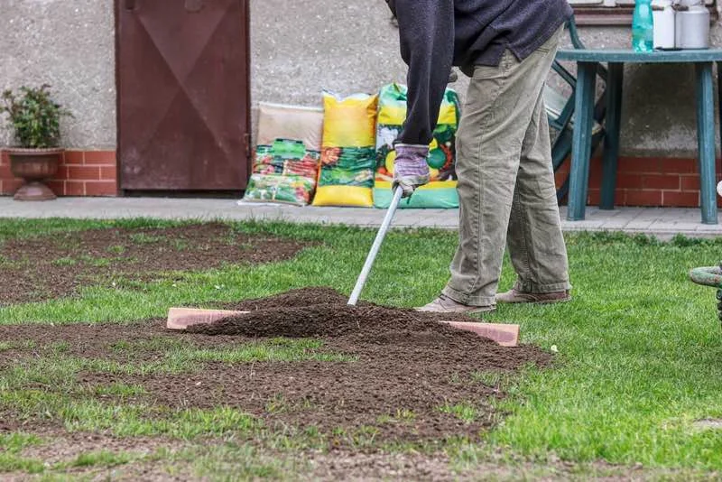 Tæt billede af en person iført handsker, der bruger en rive til at fordele jord på en græsplæne foran et hus; poser med havejord og et bord i baggrunden