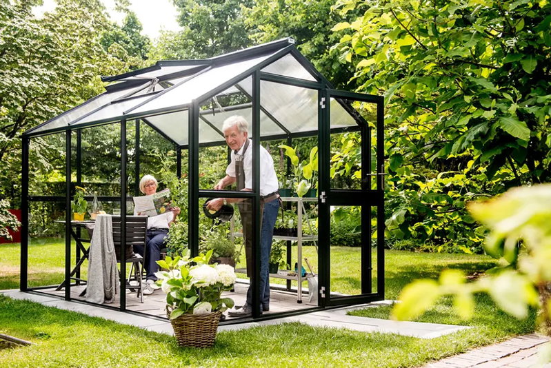 Sort drivhus i haven med ældre par, mand vander planter og kvinde læser avis Sort aluminium drivhus med glas i en grøn have; ældre mand står og vander planter inde i drivhuset, mens en ældre kvinde sidder ved et bord og læser avis. Potteplanter, haveudstyr og bænk synlige omkring dem.