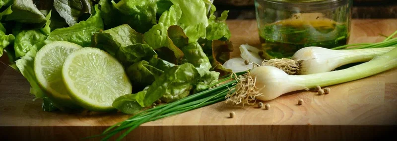 Friske salatingredienser: lime, salatblade, forårsløg og purløg på skærebræt med olivenolie Skærebræt med friske salatblade, to limebåde, purløg, forårsløg med rødder og et glas olivenolie