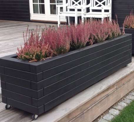 Lang sort plantekasse på hjul til terrasse Lang sort plantekasse med lyng på træterrasse