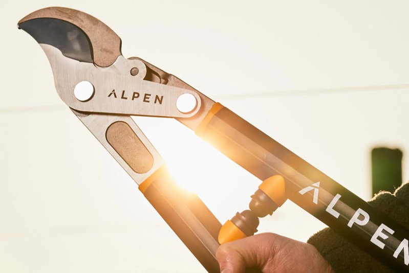 Alpen grensaks med sorte og orange håndtag og anti-slip greb Nærbillede af Alpen grensaks i brug med stålskær og sort-orange håndtag, sollys bagfra
