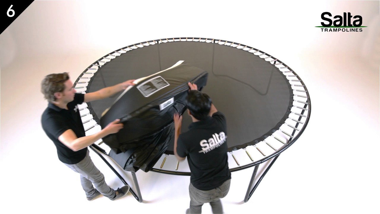 Salta Premium Black Edition trampolin Ø396 cm i sort under montage; to personer monterer den sorte kantpolstring og sikkerhedsnettet på trampolinen med sortlakerede ben og springmåtte i centrum