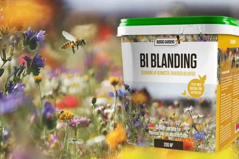 Spand med 'Bi blanding' frømix fra Nordic Gardens foran farverig blomstereng med bi i flugt, egnet til nektar- og pollenplanter