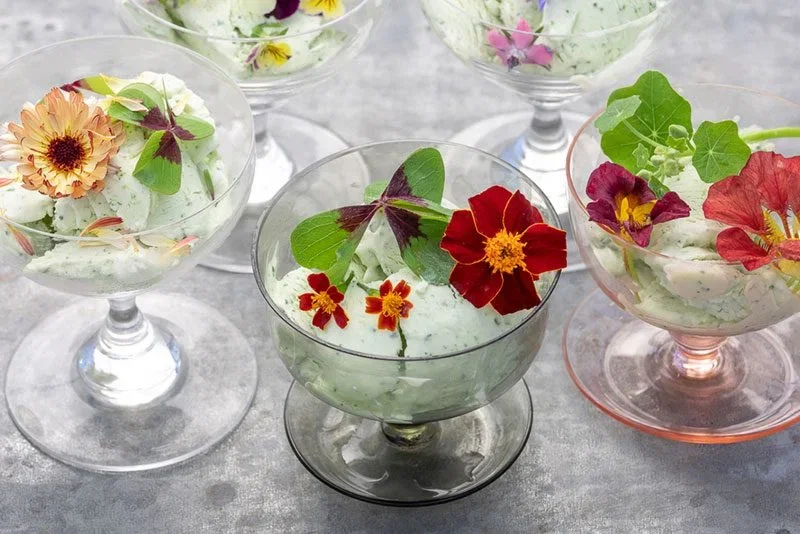 Glasdessertskåle med grøn urteis eller mousse pyntet med forskellige spiselige blomster og grønne blade på en grå bordplade