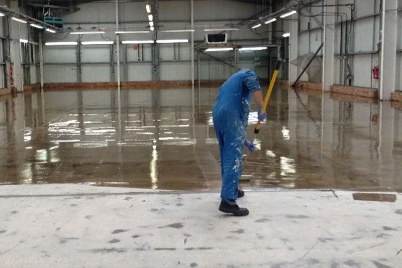 Person i blå overalls påfører gulvbelægning i stor lagerhal Person i blå overalls ruller epoxy/gulvbelægning på en poleret betonflade inde i en tom lagerhal