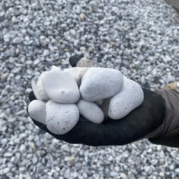 GreenBio hvide pyntesten 16–32 mm – dekorative og robuste nøddesten til indkørsel og dræn Hånd med arbejdshandske holder flere hvide pyntesten 16–32 mm fra GreenBio; runde, naturlige singels på baggrund af løst lag sten, velegnet til indkørsel, have og dræn