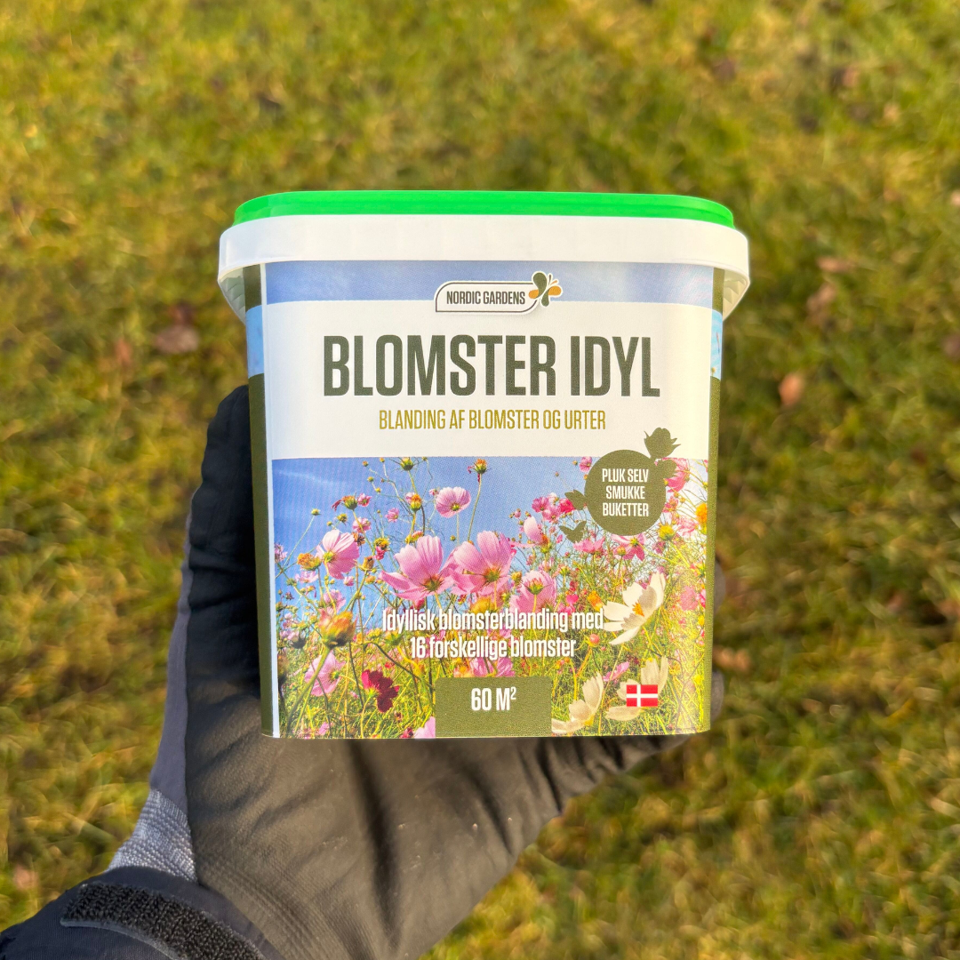 Bøtte med Nordic Gardens 'Blomster Idyl' frøblanding til 60 m², etikette med farverigt blomsterbillede. Blandingen indeholder 16 etårige blomster og urter, velegnet til havens blomsterstriber, pluk-selv buketter og som fødekilde for bier og sommerfugle.