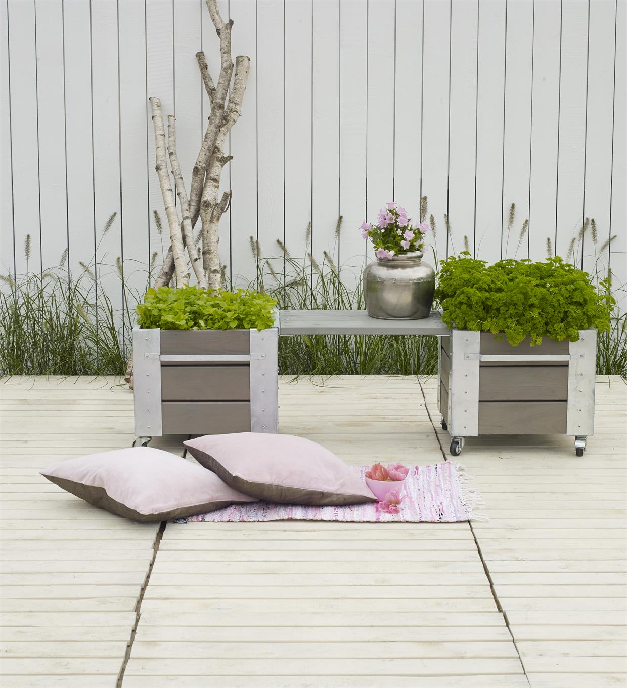 Lys træterrasse med plantekasser, blomster og puder på tæppe