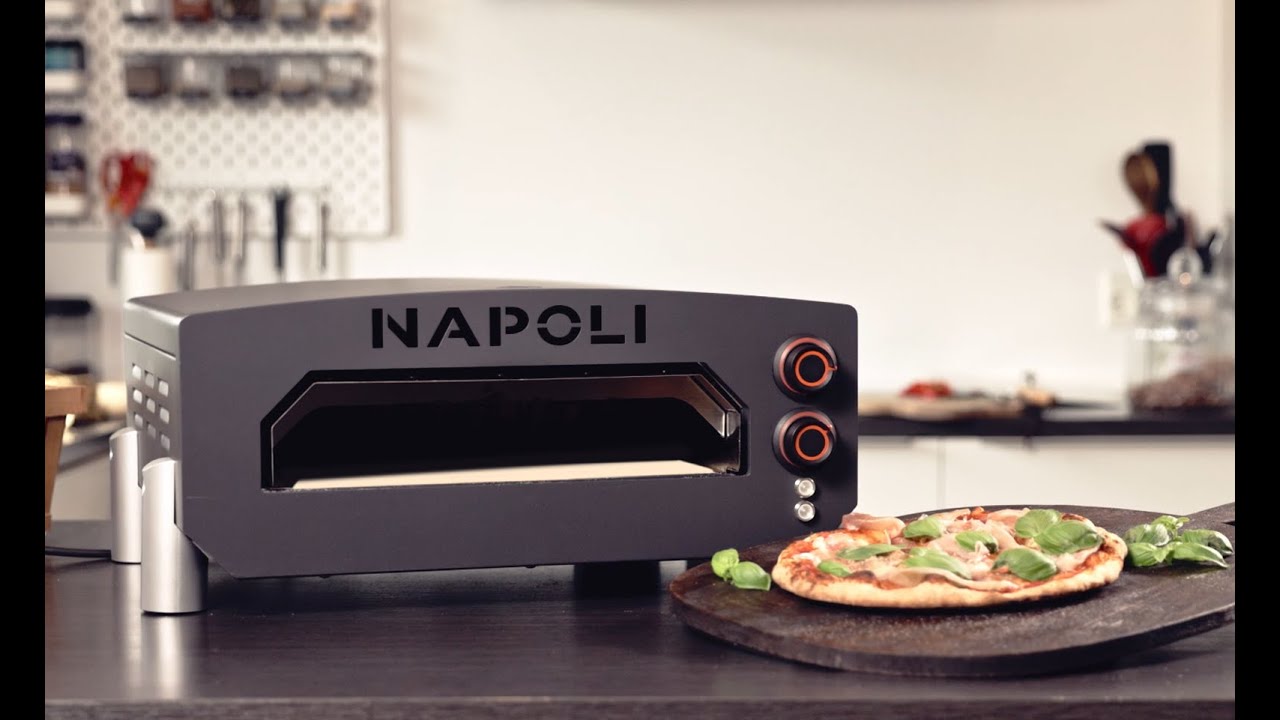 NAPOLI 13” elektrisk pizzaovn på bord med pizza på pizzasten i forgrunden, tydeligt frontvindue og betjeningsknapper. Produktpakke indeholder ovn samt tilbehør: cover, pizzaspade og termometer, egnet til hurtig opvarmning op til 400°C.