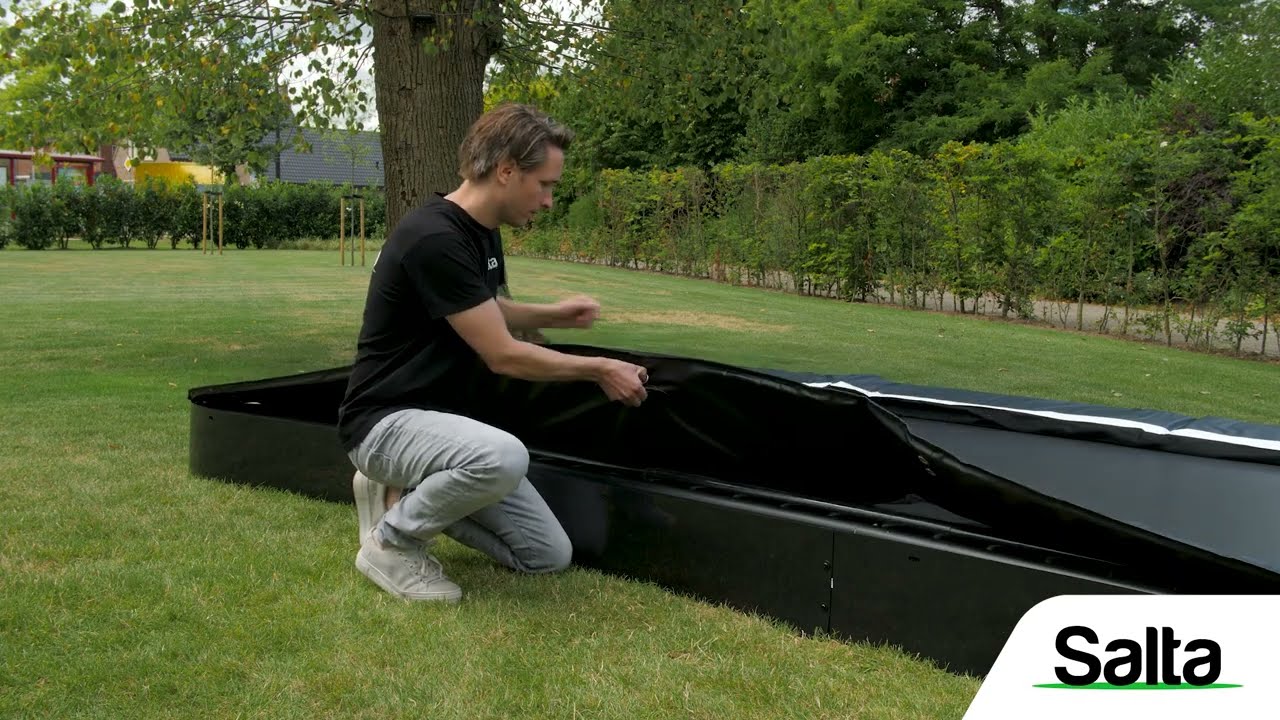 Person monterer Salta Trampolin Comfort Edition Inground Ø366 cm i sort i en nedgravet ramme, viser trampsikker kant og opslag til sikkerhedsnet – Salta
